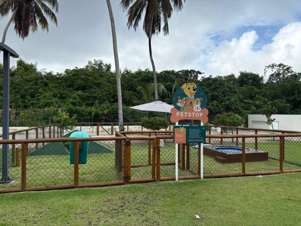 Parque para cães PetStop com cerca de madeira, gramado, rampa verde, lixeira azul e uma pequena piscina redonda. Placa com desenho de cães e o nome PetStop Dog Park