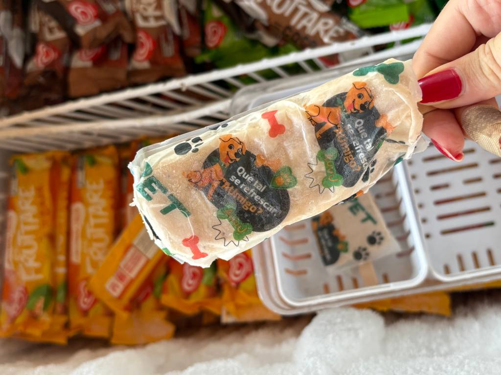 Mão feminina com unhas vermelhas segurando um picolé para pets, com embalagem transparente e desenhos de cachorro e ossos. O picolé está sendo retirado de um freezer com outros picolés e sorvetes ao fundo