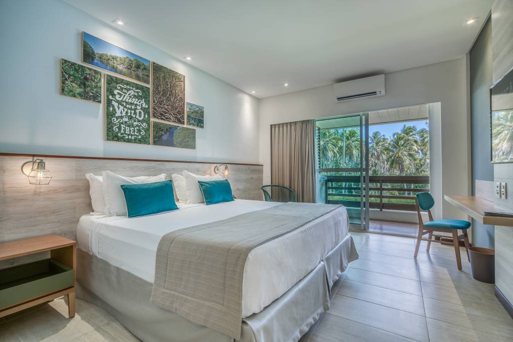 Quarto de hotel com cama de casal, lençóis brancos e almofadas turquesa. Na parede, quadros com paisagens naturais e a frase All things wild and free. Uma varanda com vista para coqueiros e um céu azul claro. À direita, uma escrivaninha de madeira com uma cadeira turquesa e um cesto de lixo. À esquerda, uma mesa de cabeceira de madeira com uma gaveta verde.