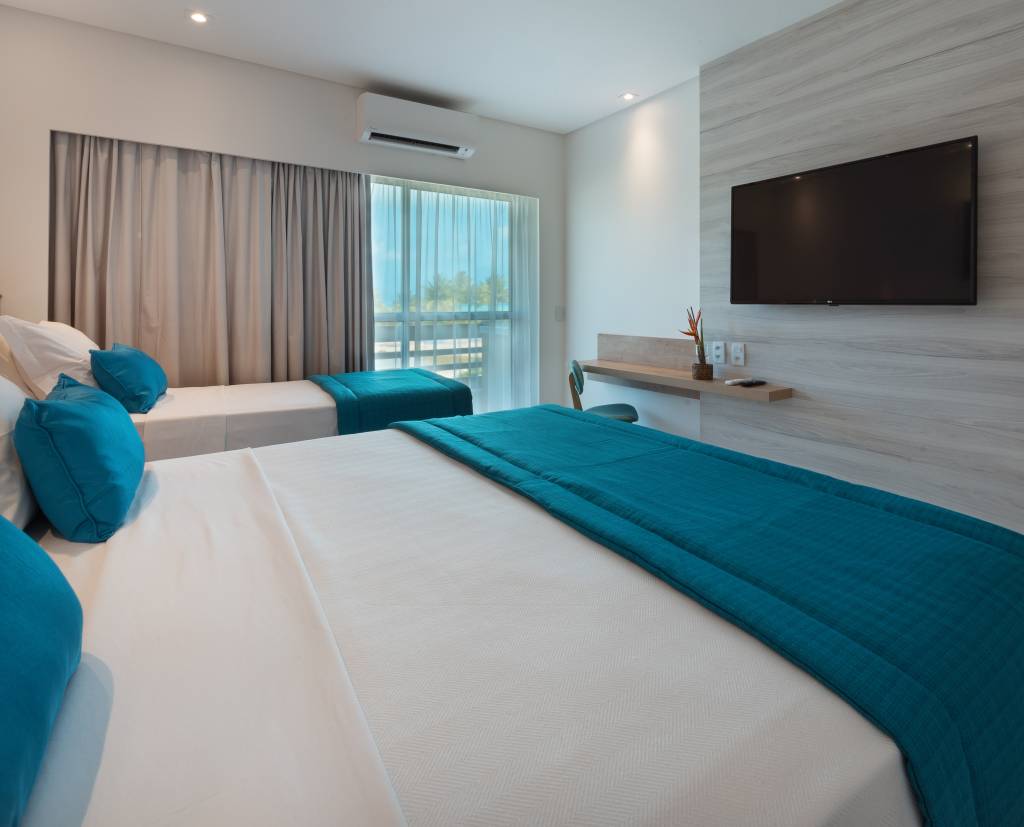 Quarto de hotel moderno com duas camas, uma de casal e uma de solteiro, ambas com lençóis brancos e mantas e almofadas turquesa. Uma TV preta está fixada na parede de madeira clara, acima de uma prateleira com um vaso de flor e um controle remoto. Uma janela com cortinas claras revela a vista para o mar