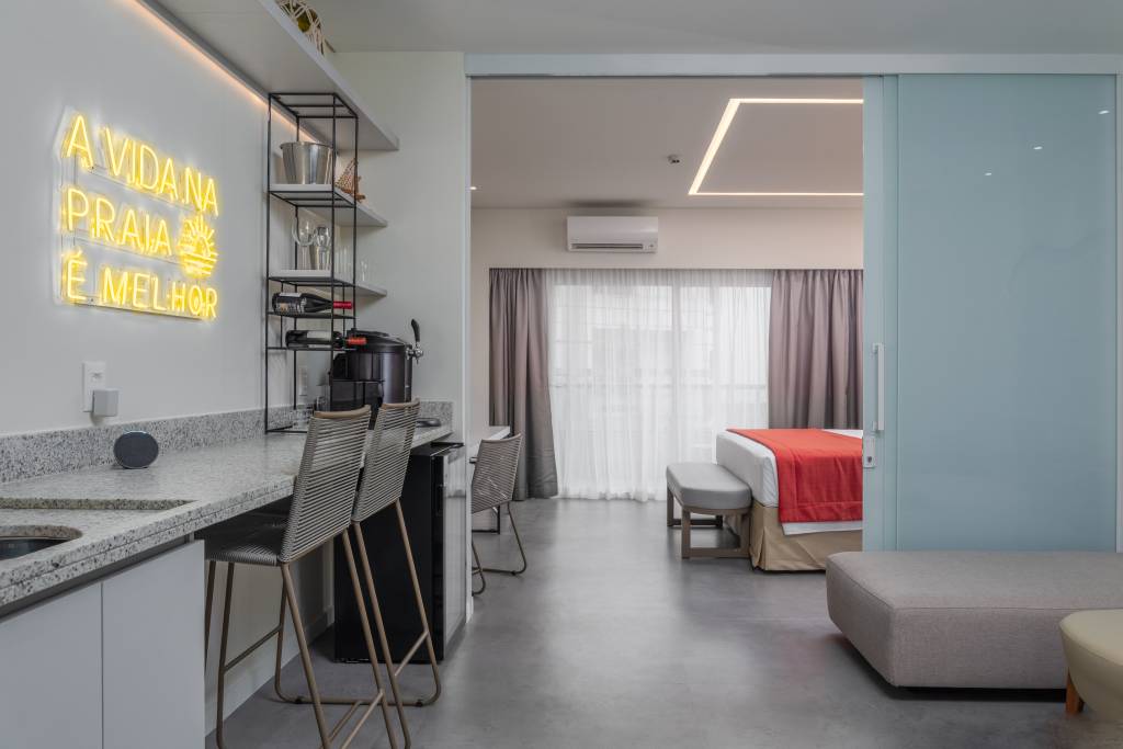 Ambiente moderno com balcão de granito, banquetas e um letreiro neon amarelo A VIDA NA PRAIA É MELHOR. Ao fundo, um quarto com cama e colcha laranja, visível através de uma porta de correr de vidro fosco