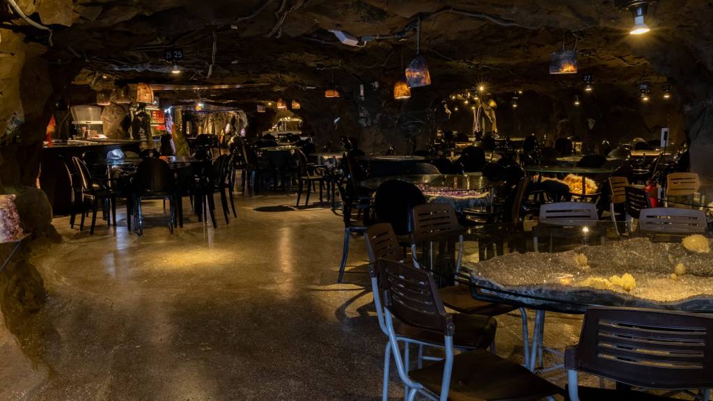 Salão de restaurante escuro com teto e paredes rochosas, mesas e cadeiras escuras, e iluminação amarelada de luminárias pendentes e embutidas. Uma mesa à direita exibe uma grande formação de cristais