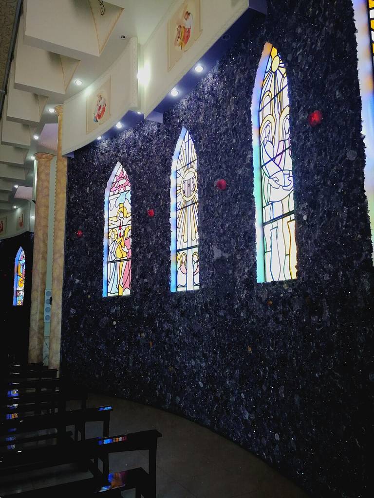 Interior de uma igreja com parede curva de pedras escuras e três vitrais coloridos em arco, iluminados. Bancos de madeira escuros em primeiro plano, e teto branco com luminárias e quadros religiosos ao fundo
