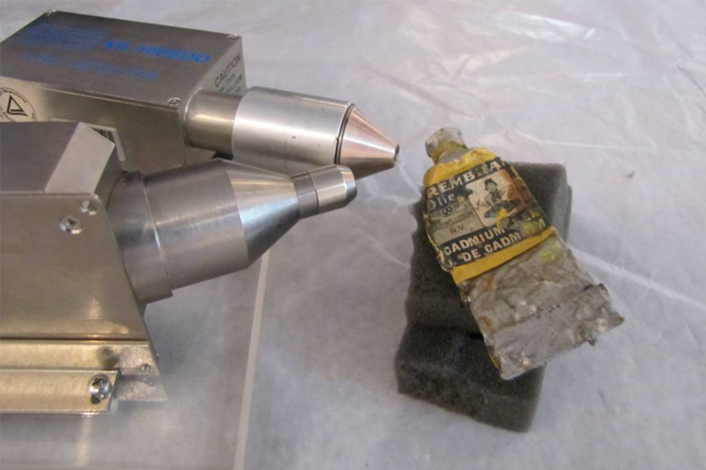 Equipamento de análise metálico com dois bicos cônicos apontados para um tubo de tinta a óleo amassado, com rótulo amarelado Rembrandt Olie e Cadmium sobre uma esponja cinza, em superfície branca