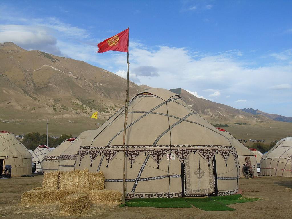 Acampamento de yurtas tradicionais no Quirguistão, com uma bandeira vermelha hasteada em frente a uma tenda decorada. Montanhas áridas e um céu azul com nuvens brancas compõem o cenário, com fardos de feno no chão.