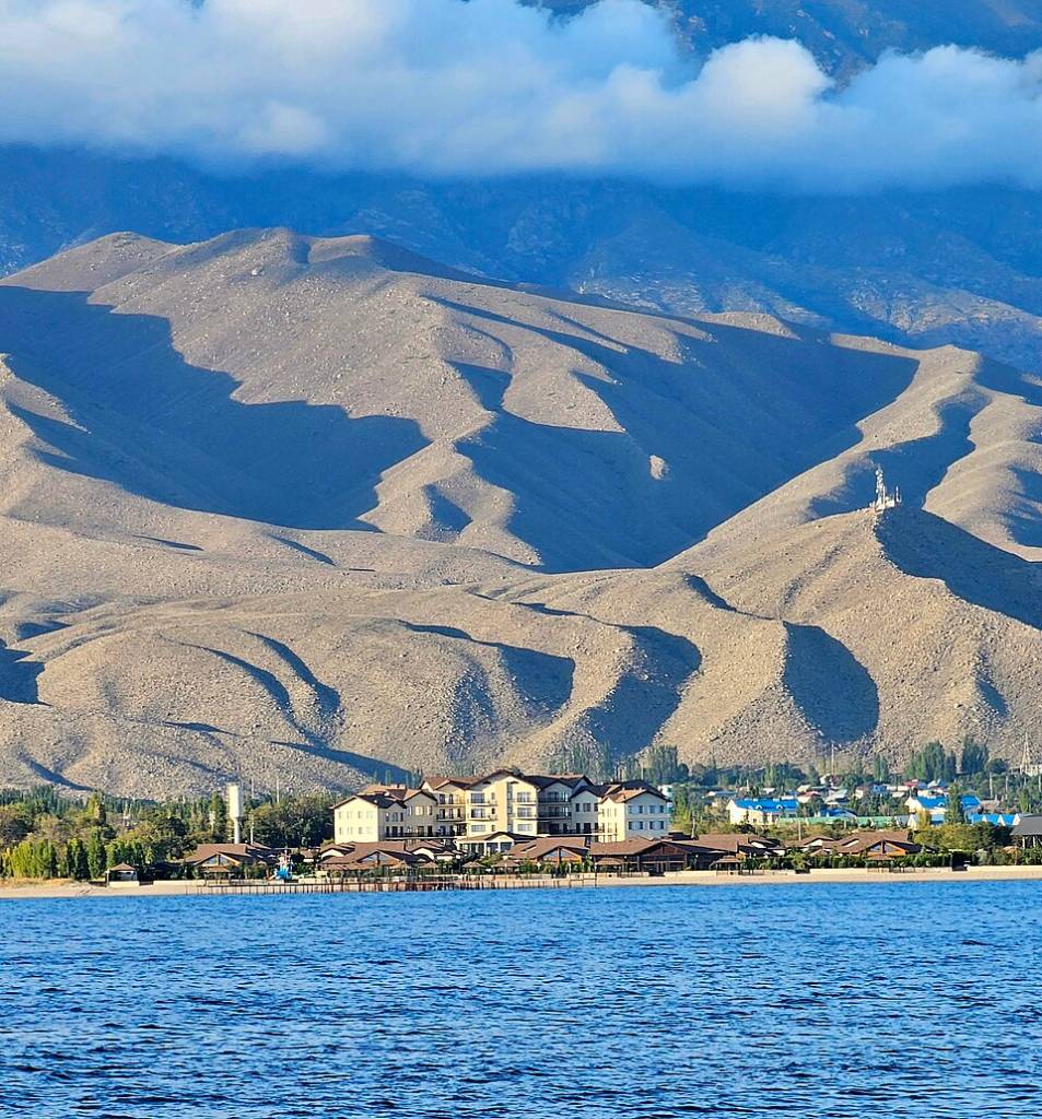 Um lago azul com ondas pequenas em primeiro plano, com um resort de vários edifícios claros e telhados escuros na margem, cercado por árvores. Ao fundo, montanhas áridas e onduladas sob um céu azul com nuvens esparsas, e uma torre de comunicação no topo de uma delas