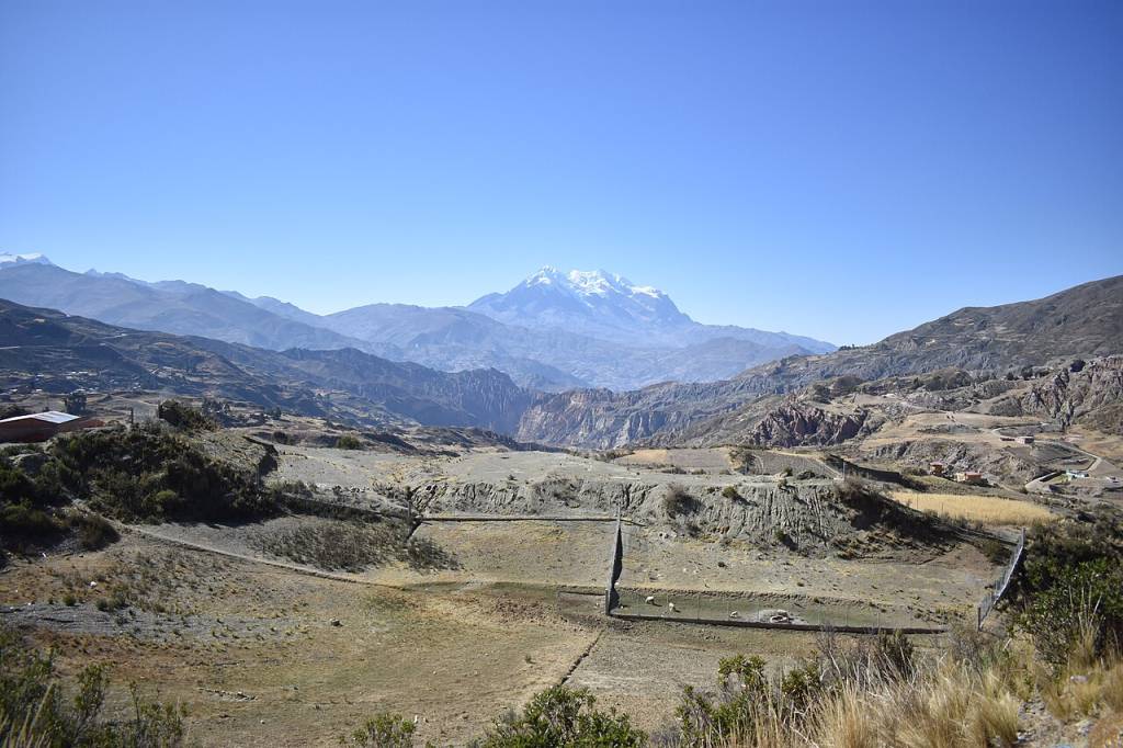 Paisagem montanhosa árida sob céu azul claro, com um pico nevado ao fundo. O primeiro plano mostra campos secos e algumas construções rurais, com vales profundos e montanhas rochosas em tons de cinza e marrom no meio