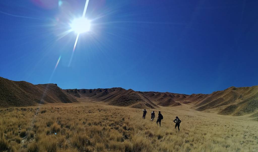 Quatro pessoas com mochilas caminham por um vale de grama seca e amarelada, sob um céu azul intenso com o sol brilhando forte no canto superior esquerdo. Ao fundo, colinas marrons e onduladas se estendem. Foto sob licença CC BY-SA 4.0