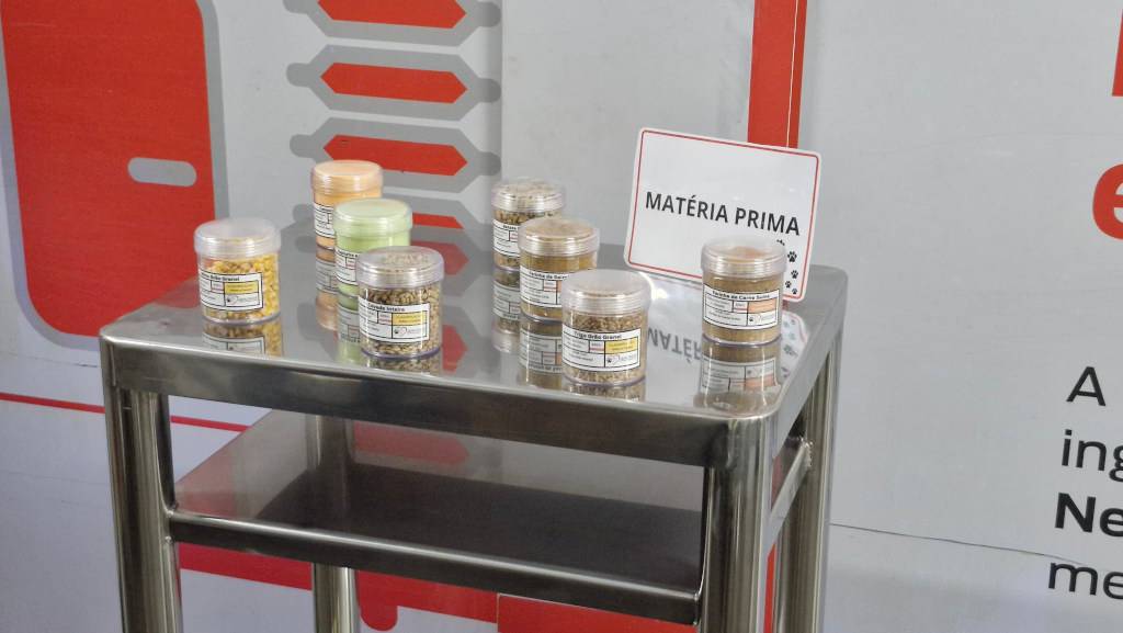 Nove potes transparentes com diferentes tipos de grãos e farinhas, identificados com rótulos brancos e amarelos, sobre uma mesa de metal. Ao fundo, um cartaz branco com a frase MATÉRIA PRIMA em letras pretas e um desenho de patas de cachorro, e um painel branco com detalhes em vermelho e cinza
