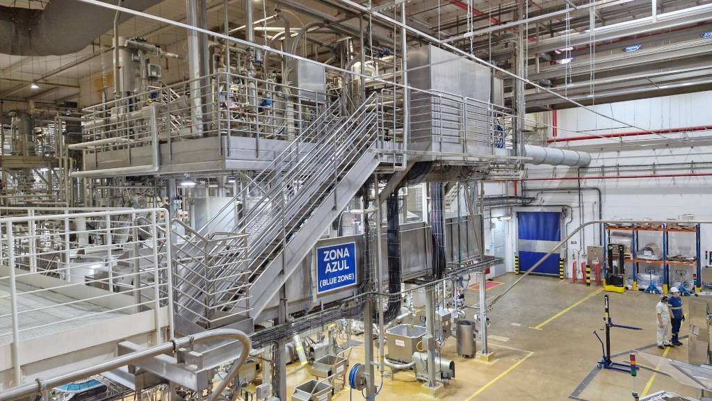 Interior de uma fábrica moderna com equipamentos industriais de aço inoxidável, tubulações e plataformas elevadas. Uma escada metálica central leva a um nível superior. Um letreiro azul na estrutura indica ZONA AZUL (BLUE ZONE). No chão, dois trabalhadores com capacetes e uniformes brancos e azuis observam o ambiente. O piso é amarelo com faixas de segurança, e ao fundo há uma porta azul e prateleiras com materiais