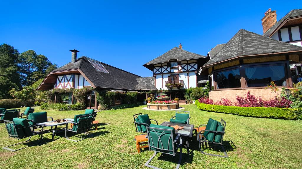 Jardim amplo com gramado verde e três conjuntos de mesas e cadeiras escuras com almofadas verdes. Ao fundo, três casas em estilo enxaimel com telhados escuros e paredes brancas, cercadas por arbustos e árvores, sob céu azul claro
