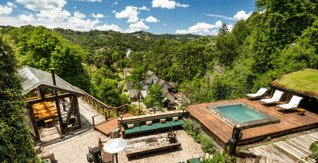 Vista aérea de um resort em meio à natureza, com piscina, espreguiçadeiras, área de convivência com sofás verdes e uma churrasqueira coberta. Ao fundo, casas e montanhas arborizadas sob céu azul com nuvens brancas
