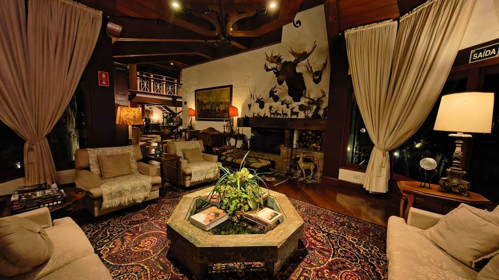 Sala de estar rústica com teto de madeira escura, lareira de pedra, e paredes brancas decoradas com cabeças de animais empalhados. Sofás e poltronas bege, tapete persa vermelho, mesa de centro octogonal com plantas e livros, e cortinas claras. Uma placa SAÍDA é visível à direita