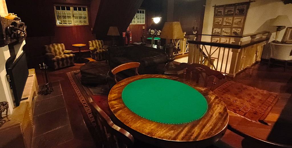 Sala de estar com mesa redonda de madeira e centro verde, cadeiras, sofás estampados, lareira, tapetes e escada com corrimão de madeira. Paredes em tons de vinho e iluminação indireta criam um ambiente acolhedor
