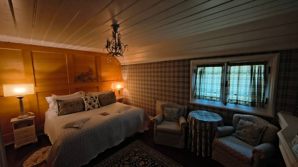 Quarto aconchegante com cama de casal, lençóis brancos e almofadas decoradas. À esquerda, uma mesa de cabeceira branca com abajur aceso. A parede da cabeceira é de madeira clara, e as outras têm papel de parede xadrez. Um lustre escuro pende do teto. À direita, duas poltronas xadrez e uma mesa redonda com toalha xadrez, perto de uma janela com cortinas. O ambiente é iluminado por luzes quentes
