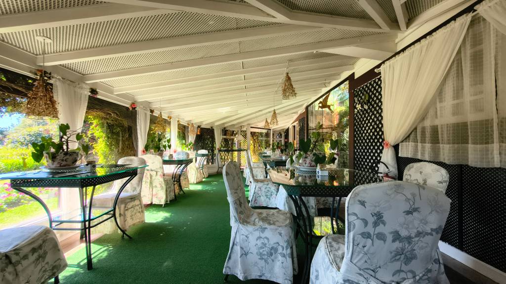 Varanda de restaurante com teto branco ripado, piso verde, mesas redondas de vidro e cadeiras com capas florais claras. Cortinas brancas translúcidas adornam as janelas, revelando folhagens verdes e flores coloridas ao fundo. Luminárias pendentes de palha e decorações de pássaros dourados complementam o ambiente.