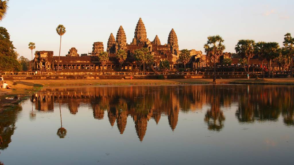 O complexo de Angkor Wat, no Camboja, com suas torres cônicas e construções de pedra, reflete-se em um lago calmo ao pôr do sol. Palmeiras e árvores emolduram o templo, e algumas pessoas caminham ou sentam na margem.