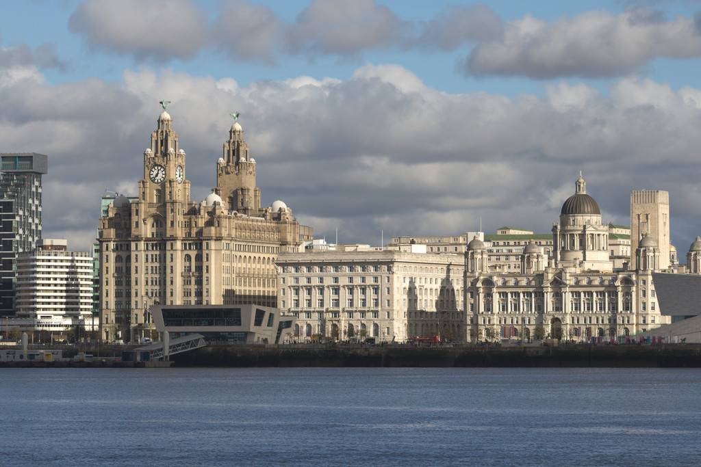 Vista panorâmica de Liverpool com o Rio Mersey em primeiro plano e edifícios históricos ao fundo, incluindo o Royal Liver Building com suas torres e relógios, o Cunard Building e o Port of Liverpool Building, sob um céu parcialmente nublado