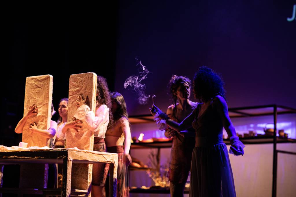 Seis mulheres em cena teatral com iluminação roxa. Duas seguram placas retangulares de papel amassado, outras duas observam uma mulher de vestido claro que segura um objeto com fumaça. Ao fundo, uma estante com objetos e a letra J projetada