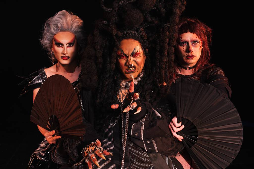 Três artistas drag com maquiagens e figurinos elaborados posam contra um fundo escuro. À esquerda, uma pessoa com peruca cinza, maquiagem dramática e batom vermelho segura um leque marrom. No centro, uma pessoa com cabelo crespo volumoso, maquiagem vermelha e piercings faciais, com expressão feroz, estende a mão com unhas longas e anéis. À direita, uma pessoa com cabelo ruivo e maquiagem gótica segura um leque preto