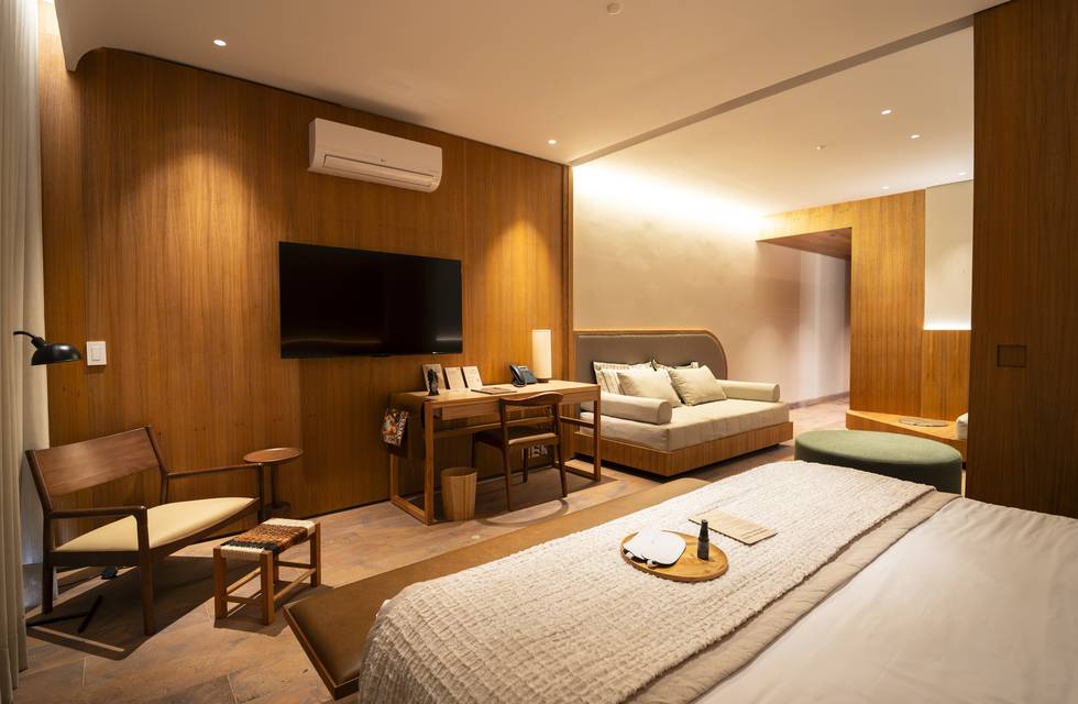 Quarto de hotel moderno com paredes de madeira clara, cama de casal com manta branca, poltrona de madeira com estofado creme, escrivaninha com cadeira, e um sofá-cama bege ao fundo