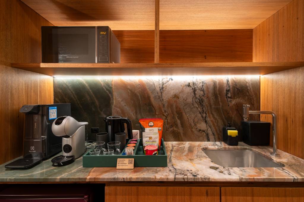 Cozinha compacta com bancada de mármore marrom e cinza, pia de inox, torneira cromada, micro-ondas Brastemp, cafeteira Dolce Gusto, chaleira elétrica, copos e sachês em bandeja verde, e prateleiras de madeira