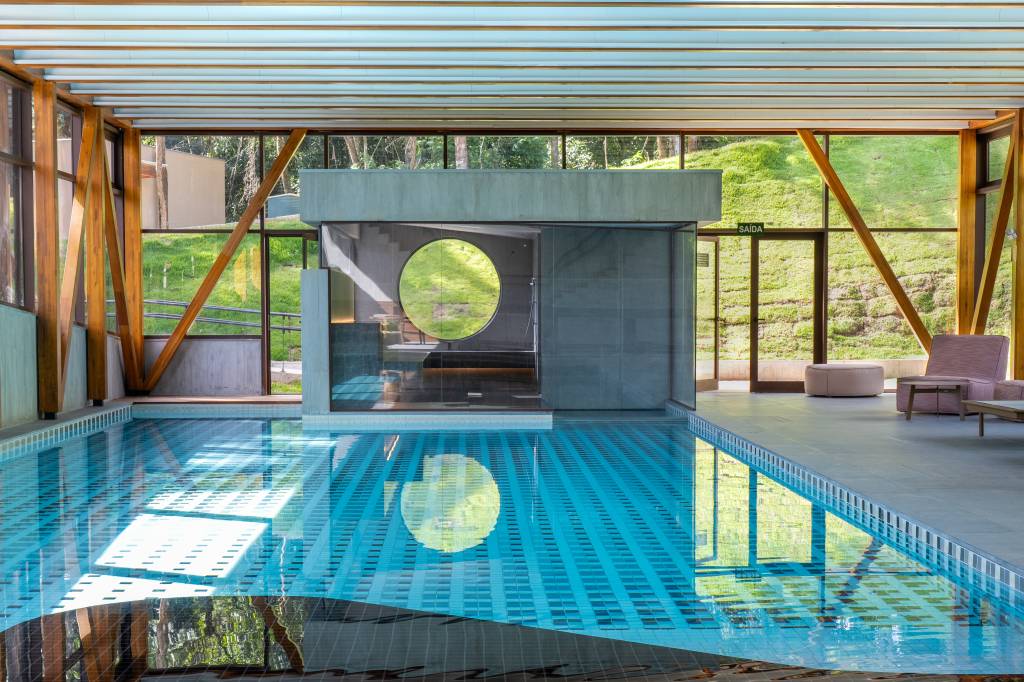 Piscina interna com água azul-clara, reflexos de luz e uma estrutura de concreto com janela circular. Ao fundo, janelas amplas revelam uma colina verde e árvores, com uma porta de saída e poltronas de descanso