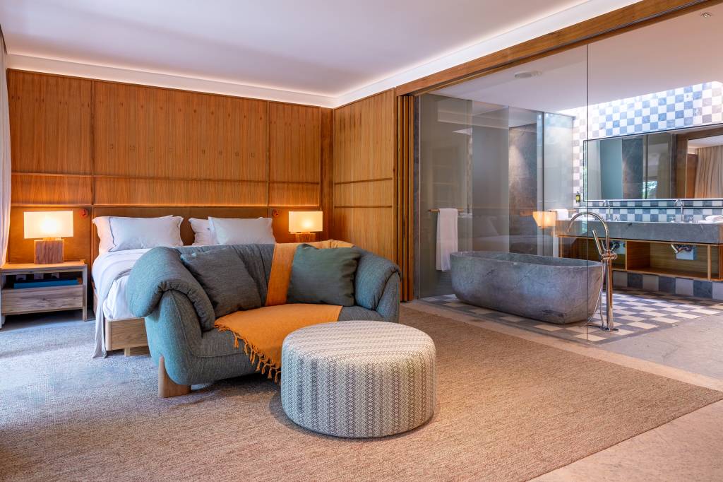Quarto de hotel luxuoso com cama de casal, sofá cinza com manta laranja, pufe estampado e banheiro integrado com banheira de pedra e pia dupla