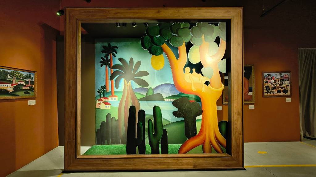 Pintura modernista colorida em moldura de madeira, com um homem deitado em uma árvore amarela, cactos, palmeiras e casas brancas em um cenário tropical com montanhas e água ao fundo