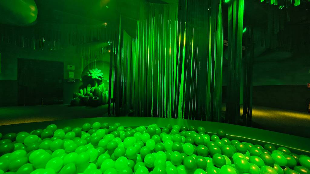 Piscina de bolinhas verde-limão em ambiente escuro com iluminação neon verde, colunas verticais e formas orgânicas brilhantes ao fundo