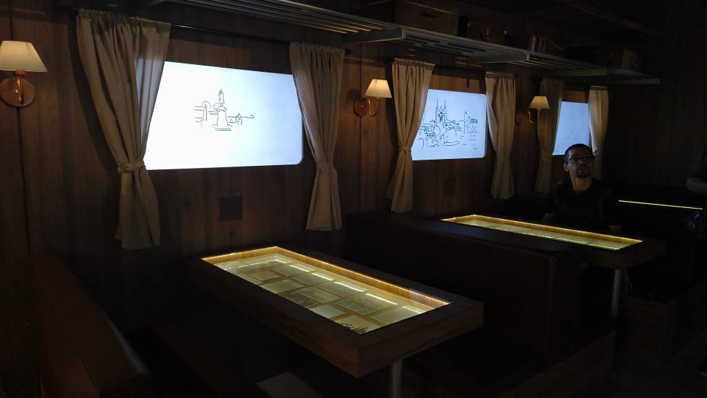 Interior de um vagão de trem escuro com paredes de madeira. Duas janelas exibem desenhos de cidades em estilo esboço. Há luminárias de parede e mesas com tampo de vidro iluminado, exibindo fotos antigas. Um homem de óculos e camiseta preta está sentado à direita