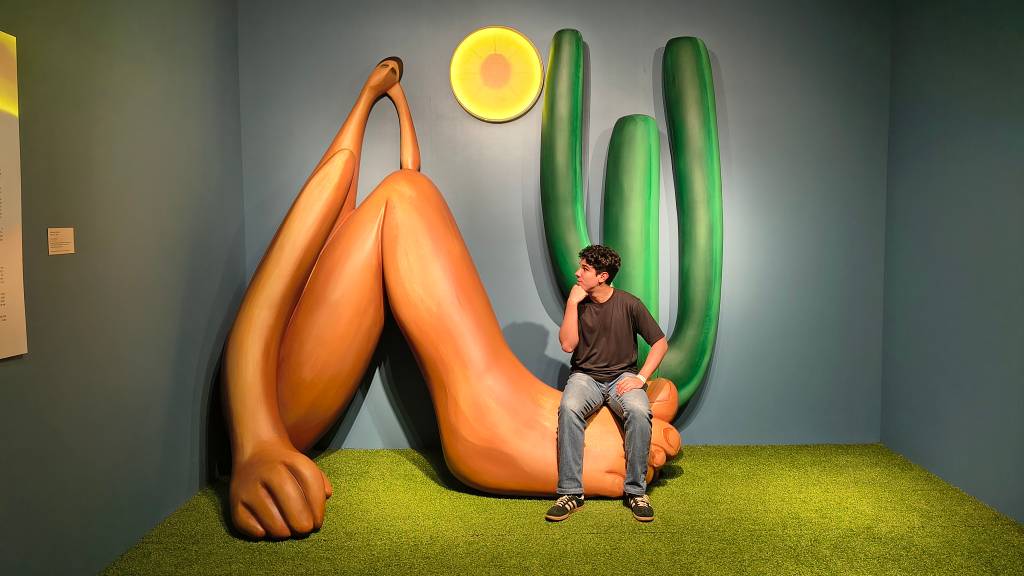 Jovem de camiseta preta e jeans sentado sobre um pé gigante de escultura cor de pele, olhando para a direita. Ao fundo, parede azul com um sol amarelo e três cactos verdes, e chão de grama sintética