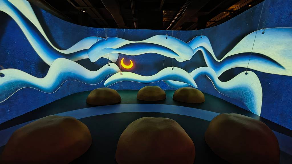 Instalação artística imersiva com projeções de ondas azuis e brancas em uma parede curva, com uma lua crescente amarela no centro. No chão, seis formas arredondadas marrons simulam pedras, criando um ambiente noturno e sereno