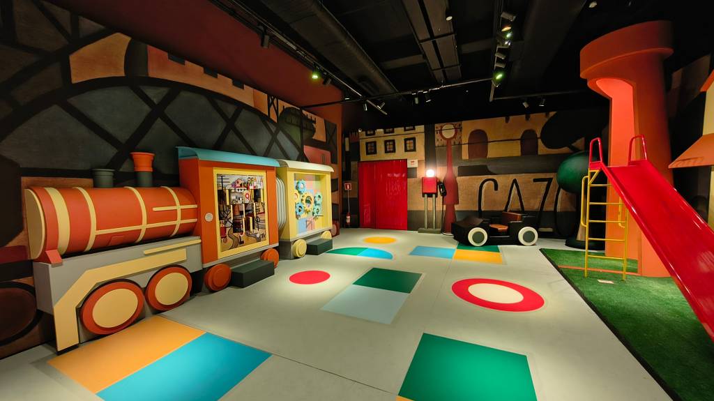 Cenário de brinquedos coloridos com um trem laranja e amarelo à esquerda, um carro preto com rodas brancas ao centro, e um escorregador vermelho com escada amarela à direita. O chão tem formas geométricas coloridas e as paredes são pintadas com elementos urbanos estilizados