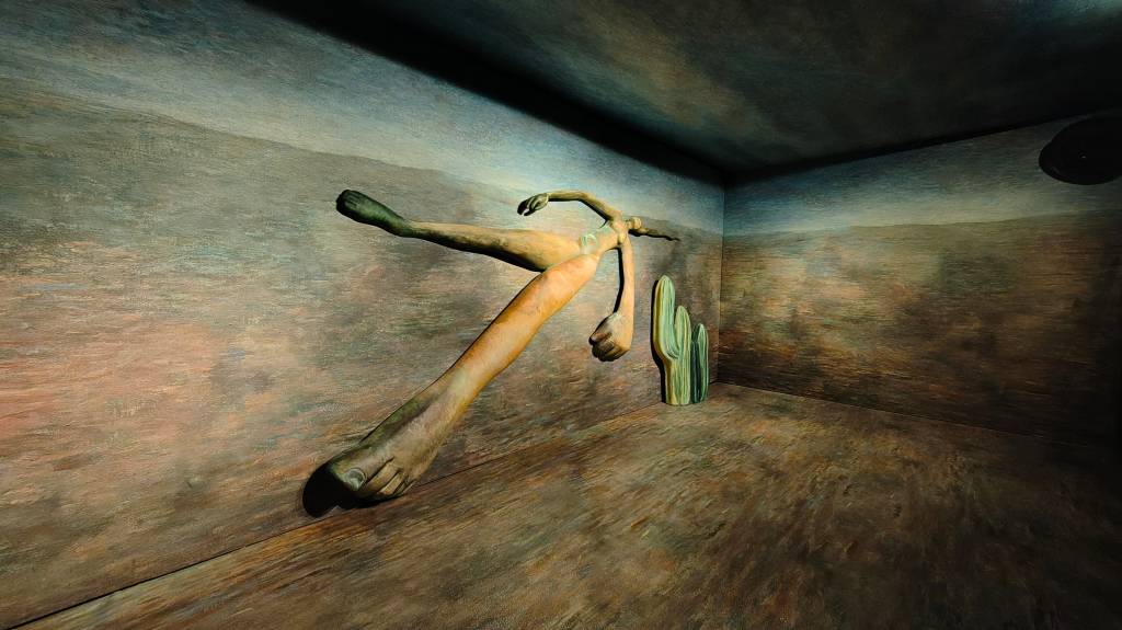 Escultura de figura humana alongada, deitada de lado, com um braço estendido e pernas esticadas, em um ambiente que simula um deserto com um cacto ao fundo