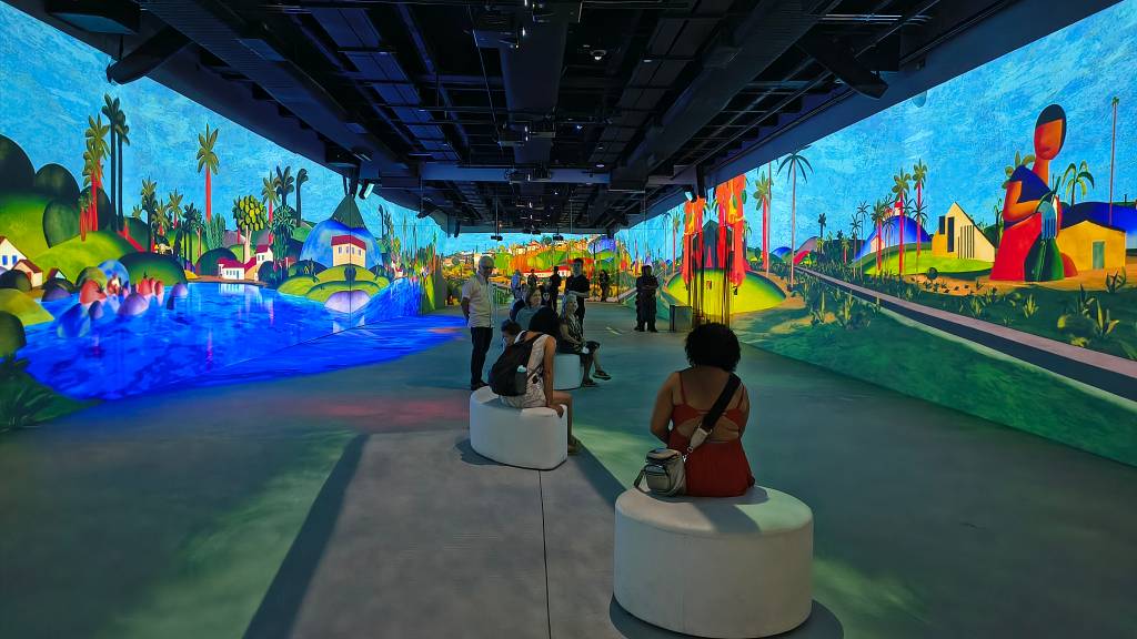 Pessoas observam uma exposição imersiva com projeções de paisagens coloridas no estilo de Tarsila do Amaral, com rios azuis, casas e coqueiros, em um ambiente escuro com bancos brancos