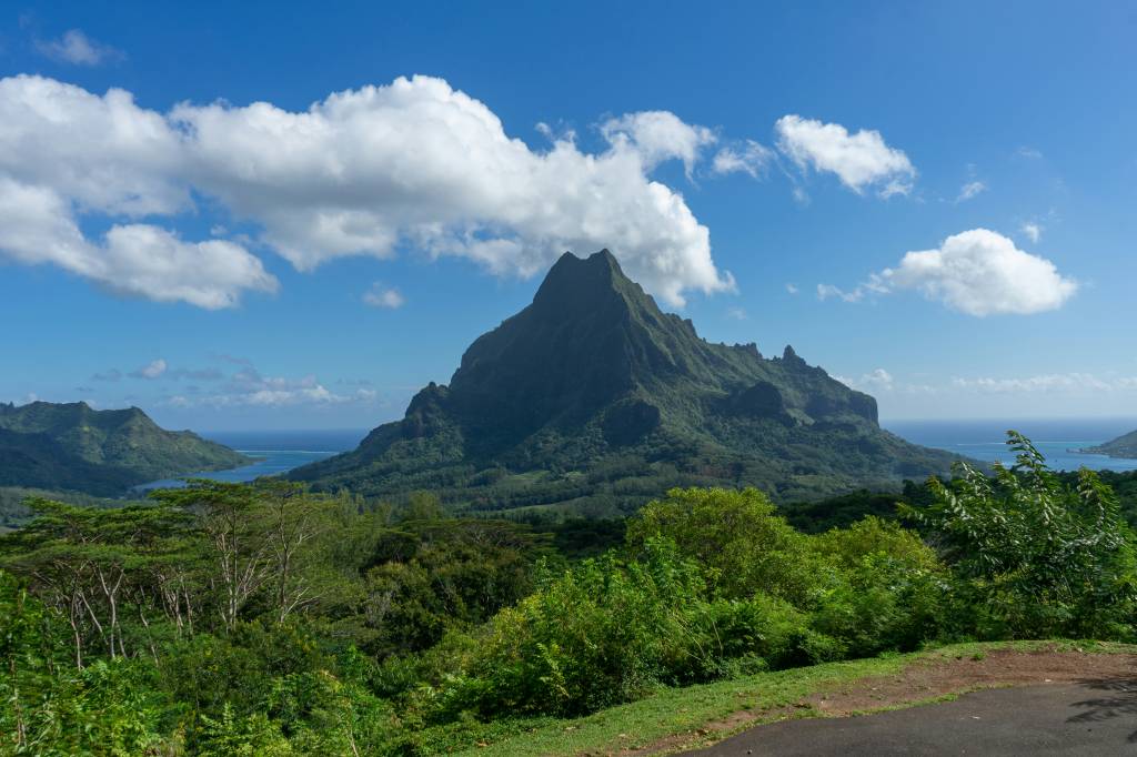 Montanha verde exuberante com picos pontiagudos sob céu azul com nuvens brancas, cercada por floresta densa e mar azul ao fundo, com uma estrada de terra no canto inferior direito