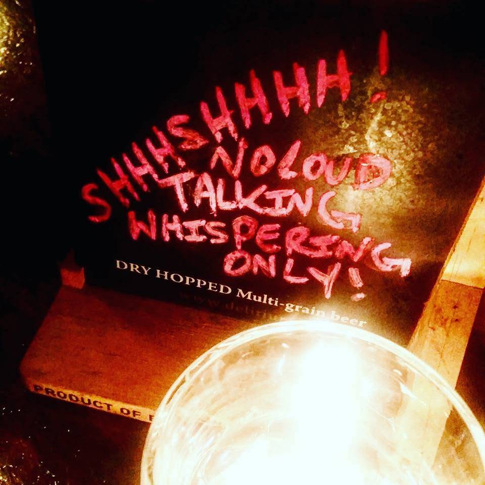 Rótulo de cerveja escura com a frase SHHHSHHHH! NO LOUD TALKING WHISPERING ONLY! em letras vermelhas e DRY HOPPED Multi-grain beer em branco. Uma vela acesa em um copo de vidro ilumina a cena, criando um ambiente intimista
