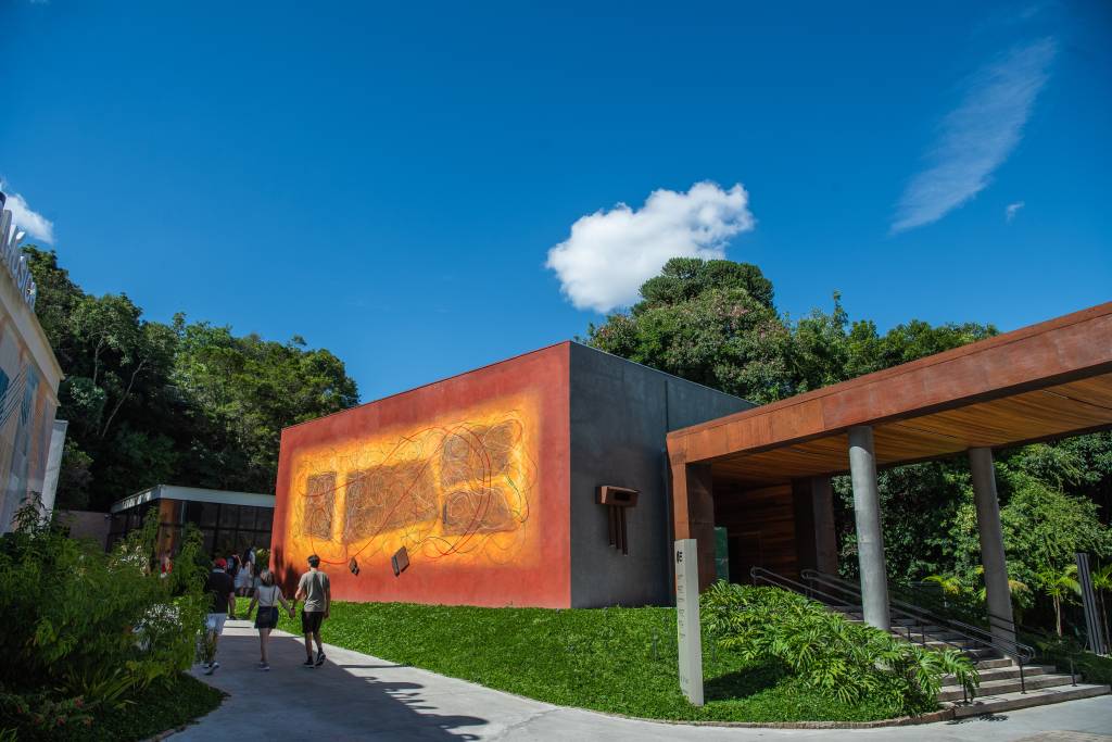 Pessoas caminham em uma trilha de cimento entre edifícios modernos e vegetação exuberante sob céu azul com nuvens. À esquerda, um prédio claro com letreiro MÚSICA. À direita, um edifício cinza com uma grande parede laranja e amarela decorada com grafismos abstratos. Mais à direita, uma estrutura de madeira e metal com pilares de concreto e escadas, cercada por plantas. O ambiente é de um parque ou centro cultural.