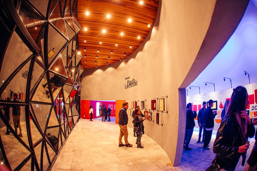 Pessoas visitam uma exposição com paredes de espelhos triangulares e uma parede branca com a inscrição Espaço Paulo Leminski e quadros pendurados. O teto é de madeira com iluminação embutida
