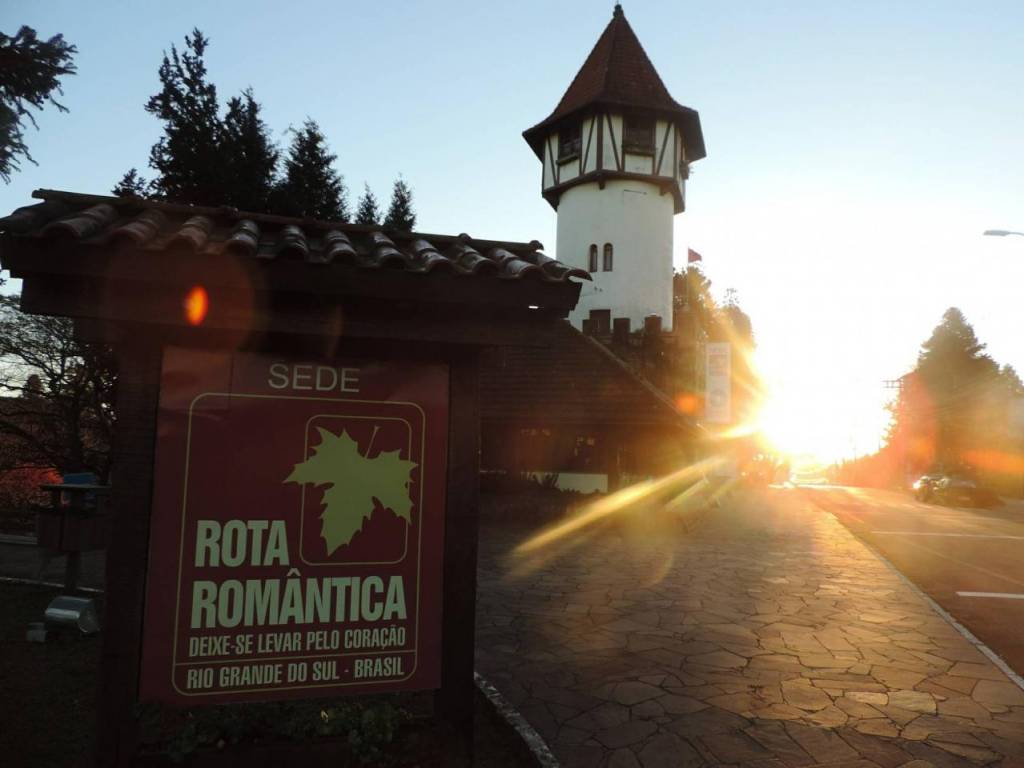Placa da Rota Romântica com folha verde e texto Deixe-se levar pelo coração Rio Grande do Sul - Brasil, em frente a um prédio com torre estilo europeu, sob o sol poente que ilumina a rua de paralelepípedos