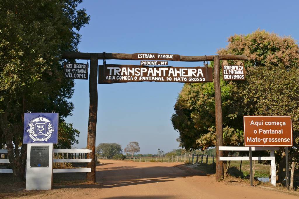 Portal de madeira rústico com placas de boas-vindas à Transpantaneira e ao Pantanal Mato-grossense em português, inglês e espanhol. À esquerda, um totem com brasão azul e placa informativa. Uma estrada de terra se estende ao fundo, ladeada por árvores e vegetação sob céu azul claro. Foto sob licença CC BY-SA 2.0