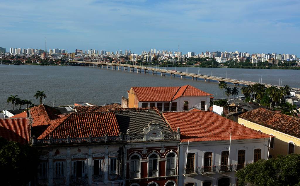 Vista aérea do centro histórico de São Luís, no Maranhão, com telhados coloniais de barro em primeiro plano, um rio largo com uma ponte longa ao centro, e a cidade com arranha-céus ao fundo, sob um céu azul claro