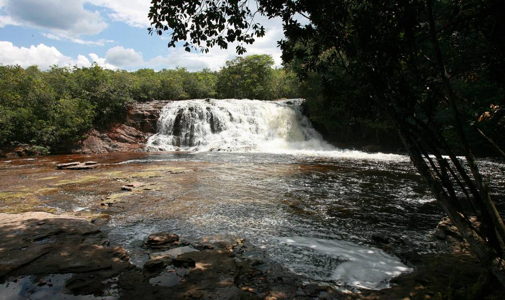 Cachoeira de águas brancas caindo sobre rochas avermelhadas, cercada por vegetação densa e árvores escuras à direita, sob céu azul com nuvens esparsas
