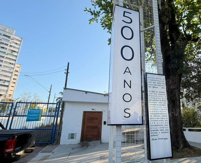 Placa vertical branca com 500 ANOS em preto e outra placa menor com texto sobre o Monumento Comemorativo aos 500 Anos do Descobrimento do Brasil, 2002, ao lado de uma árvore e um prédio alto, sob céu azul