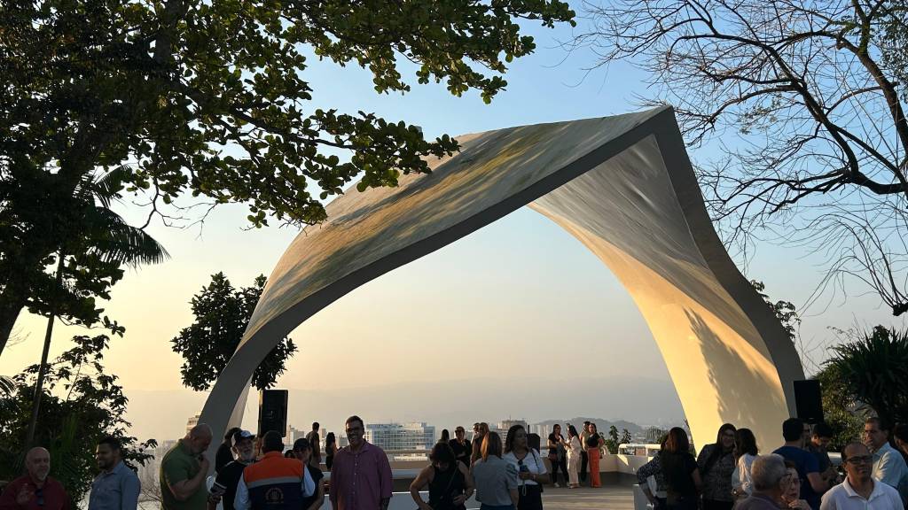 Pessoas reunidas sob um arco arquitetônico moderno, com árvores frondosas e o céu azul-claro ao fundo, em um entardecer ensolarado