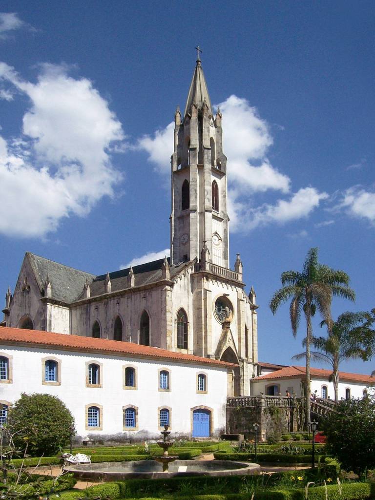 Igreja de estilo gótico com torre alta e pontiaguda, fachada clara, telhado escuro, e um edifício branco com telhado de terracota e janelas azuis em primeiro plano. Um jardim com fonte e palmeiras completa a cena sob céu azul com nuvens esparsas. Foto sob licença CC BY 3.0