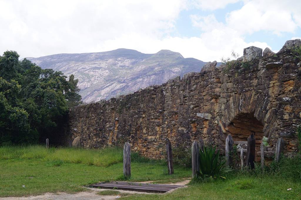 Muro de pedra antigo com um arco, em meio a gramado verde e árvores, com montanhas rochosas ao fundo sob céu nublado. Foto sob licença CC BY-SA 4.0