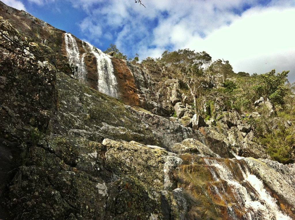 Cachoeira em montanha rochosa com vegetação esparsa e céu azul com nuvens brancas. A água desce por várias fendas na rocha, formando pequenas quedas e corredeiras. A rocha é coberta por musgo e líquen em tons de verde e marrom. Foto sob licença CC BY-SA 3.0