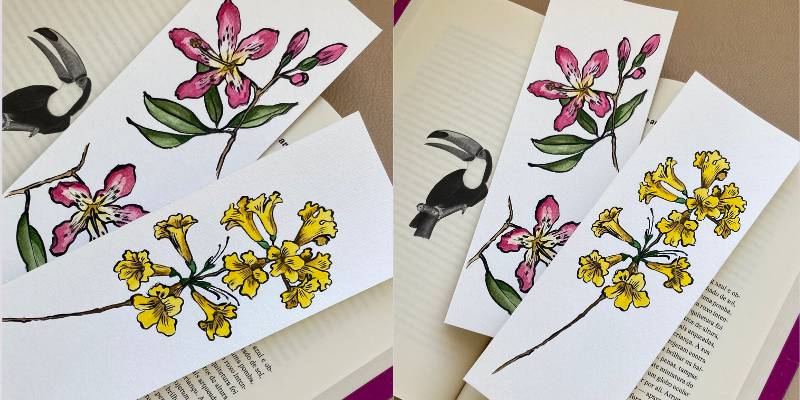 Três marcadores de página brancos com ilustrações botânicas e de animais, sobre um livro aberto. Um marcador tem um tucano preto e branco, outro tem flores cor-de-rosa com folhas verdes, e o terceiro tem flores amarelas em forma de sino com folhas verdes.